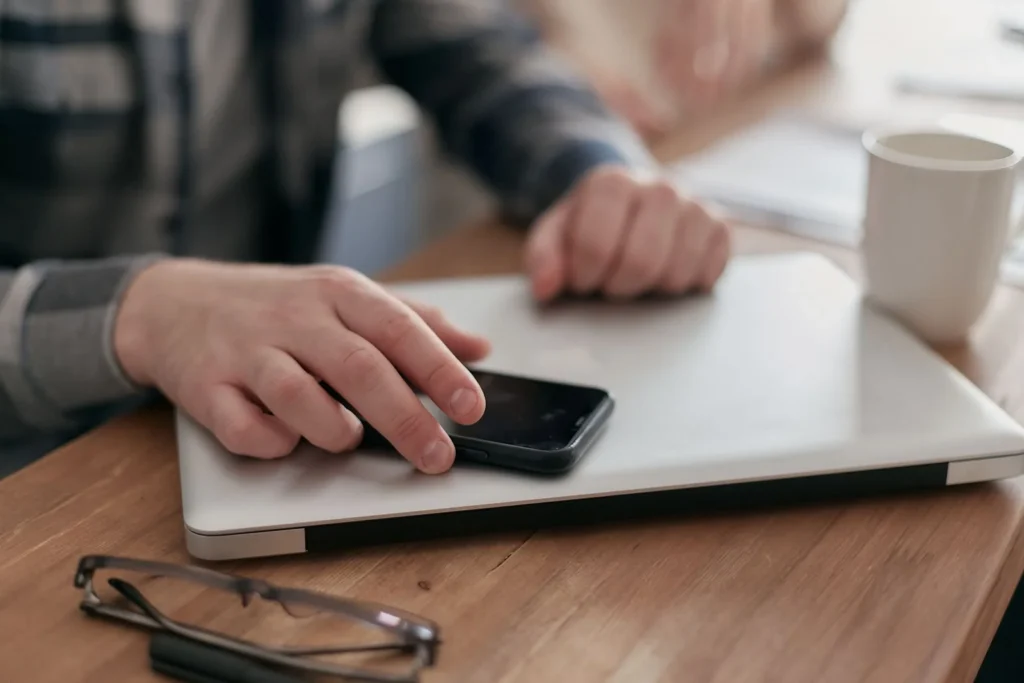 Hender med mobil og laptop ved skrivebordet