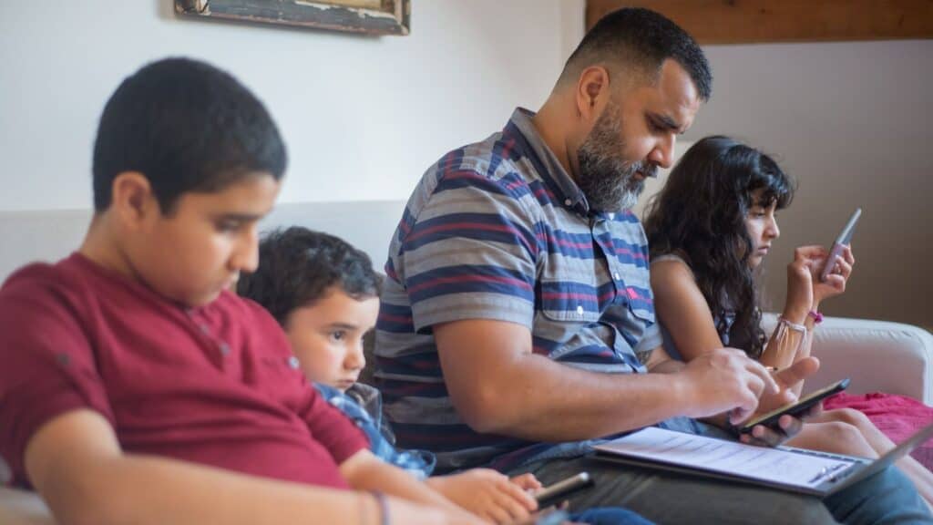 Far og tre barn sitter i sofaen og bruker hver sin mobil, nettbrett og PC