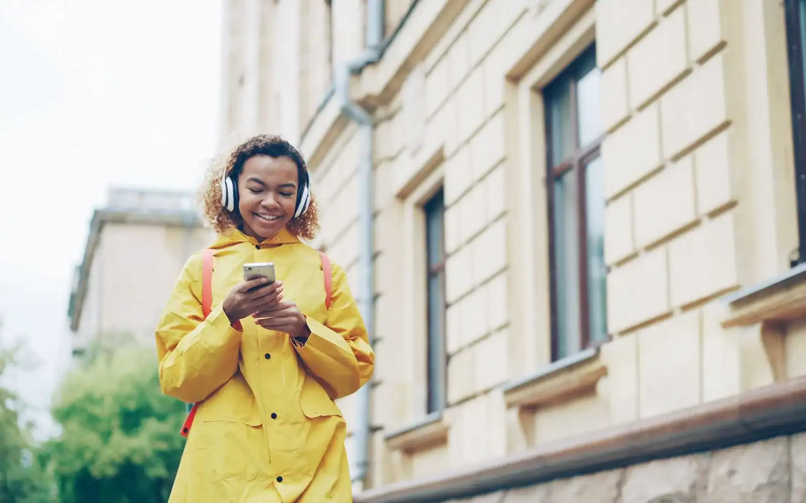 Smilende kvinne i gul regnjakke med hodetelefoner og mobil på et fortau