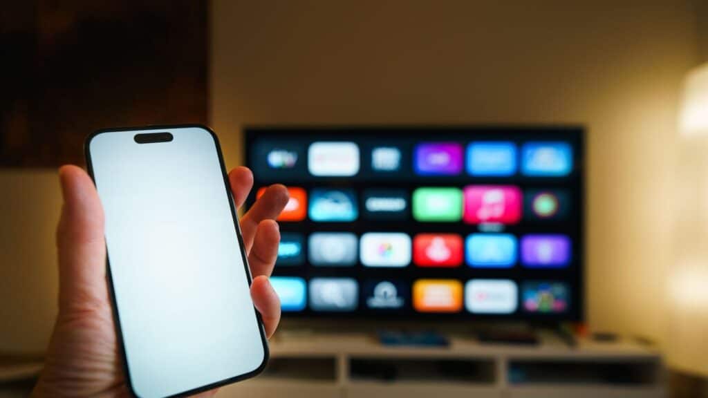 Mobiltelefon holdt foran en smart-TV som viser app-menyen med strømmetjenester