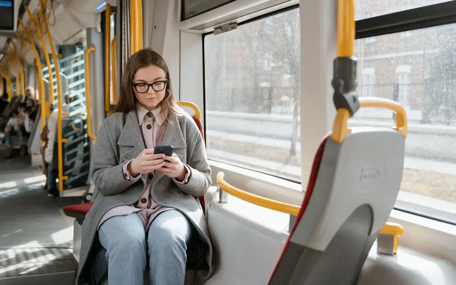 Kvinne i grå kåpe sitter på trikken og bruker mobilen