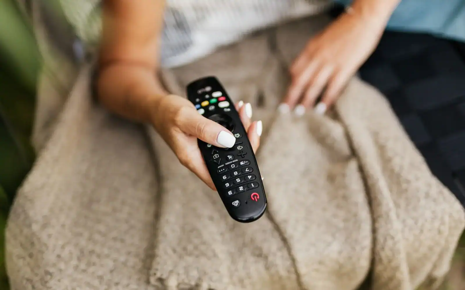 Person under et pledd holder fjernkontrollen i sofaen
